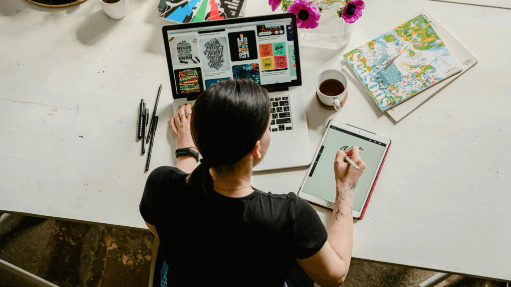 Create your brand identity How to start a clothing brand A woman is sitting at a desk, using a laptop and pad to create graphic design elements.