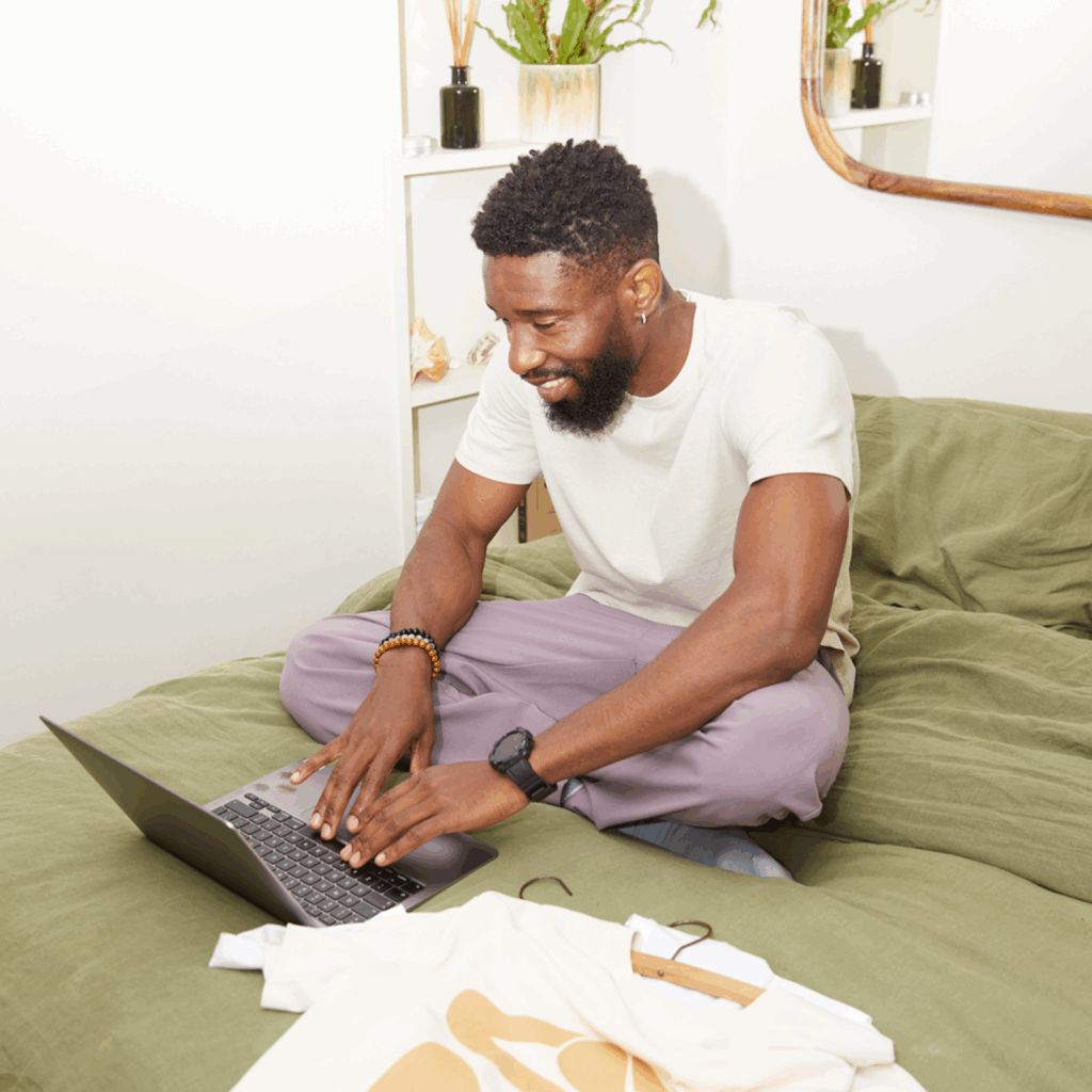 Print on Demand How to start a clothing brand A smiling man is sitting on a bed, using a laptop, with a custom t-shirt laying near the laptop.